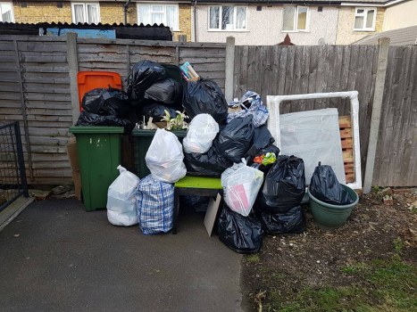 A large collection of black and white rubbish bags, some tied and others open, are piled on and around a green outdoor bench situated on a concrete driveway. The bags contain household waste and are stacked haphazardly, with some leaning against a green wheeled bin positioned to the left of the bench. Behind the pile, there is a wooden fence with vertical panels, framing the scene and separating it from adjacent residential buildings in the background. To the right of the rubbish bags, an empty white frame or panel leans against the fence, and a green plastic planter or basin is placed on the ground. The environment appears to be an outdoor area in Bounds Green, with natural daylight illuminating the scene, indicating a typical urban backyard or side alley suitable for rubbish collection and clearance services provided by Office Clearance Bounds Green, based in North London postcode areas.