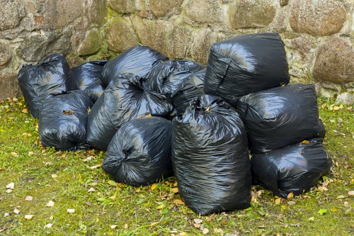 A collection of multiple black plastic rubbish bags, filled with waste materials, piled on a grassy area next to a weathered stone wall. The bags are tightly tied and stacked in a somewhat uneven manner, with some lying on their sides and others upright. The surface of the bags appears smooth and shiny, indicating they are made of standard plastic. The surrounding environment features patches of green grass with scattered small stones and fallen leaves, suggesting an outdoor setting. Behind the bags, the stone wall is constructed of irregularly shaped rocks with moss and lichen growth, adding natural texture to the background. The image, associated with rubbish removal services, highlights waste awaiting collection, possibly as part of a local clearance or refuse disposal process, relevant to companies like Office Clearance Bounds Green serving the London area near postcode N11.