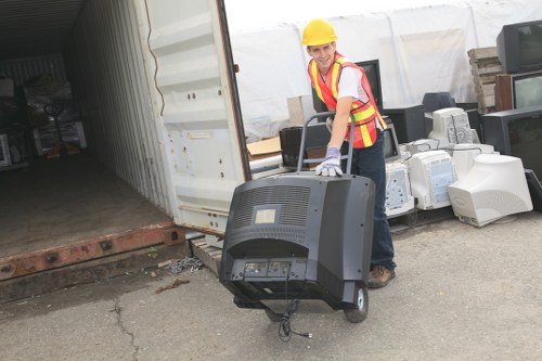 A woman wearing a yellow safety helmet, high-visibility vest, and gloves is standing outside a storage unit doorway on a paved surface, lifting and carrying a black computer tower. The storage unit has an open metal sliding door revealing shelves with various items inside, including boxes and other office equipment. To the right, several old computer monitors and electronic waste are stacked on the ground, with some leaning against the exterior of the storage unit. Behind her, there is a collection of discarded electronic devices, likely destined for rubbish removal. The scene appears to be part of an office clearance operation, with a focus on sorting and removing electronic waste, and is set in a commercial or industrial area consistent with locations in Bounds Green or nearby London suburbs. The overall environment is well-lit, with natural daylight illuminating the scene, emphasizing the professional and neutral tone of waste management activities undertaken by Office Clearance Bounds Green.