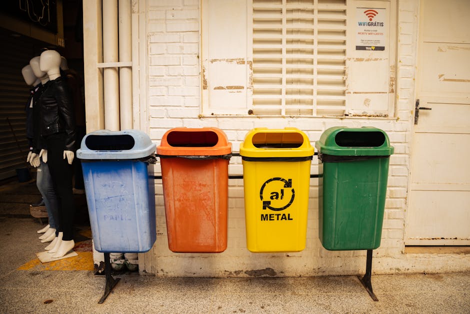 A green outdoor waste recycling bin mounted on two metal posts positioned against a yellow tiled wall. The bin features a black icon indicating it is for general waste disposal, with a small red indic