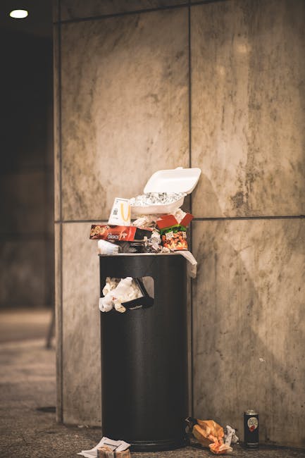 A black cylindrical rubbish bin made of metal with a closed lid is positioned against a large, light brown tiled wall in an indoor setting. The bin is overflowing with various discarded takeaway food