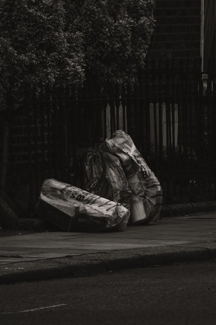 Three large plastic bags filled with miscellaneous waste materials are positioned on the edge of a paved sidewalk next to a black metal fence. The bags appear to contain cardboard, paper, and other ho