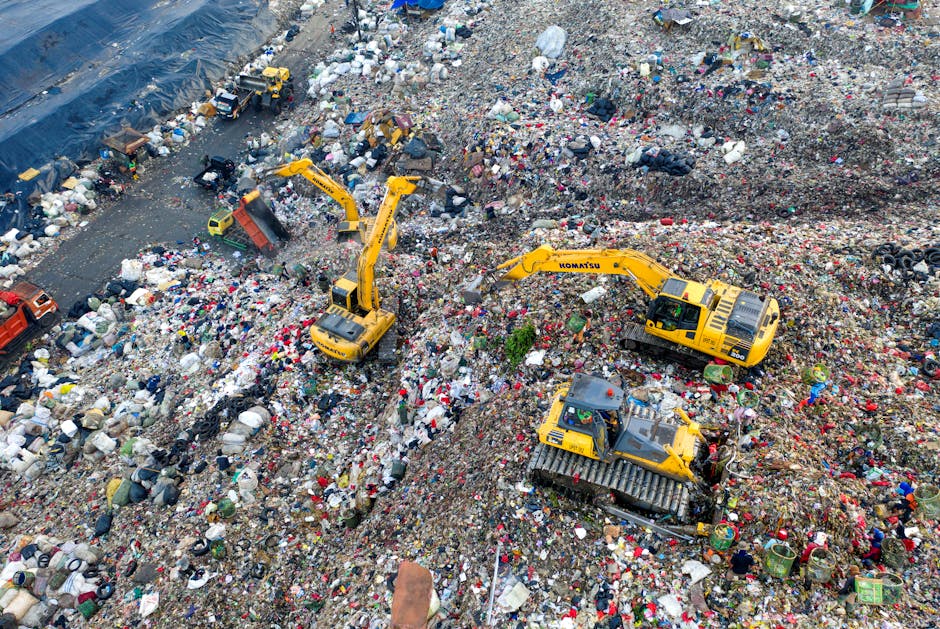 A close-up view of mixed rubbish piled on the ground outdoors, comprising black plastic garbage bags, discarded cardboard boxes, a large yellow plastic container, an old beige car seat, and pieces of 